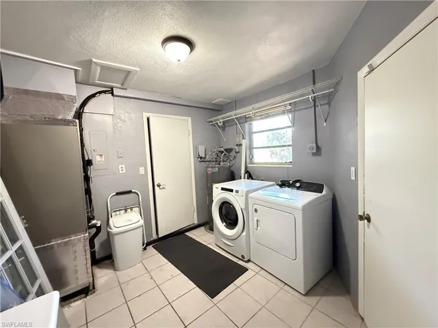 Laundry area with heating unit, a textured ceiling, washer and clothes dryer, light tile patterned floors, and electric panel