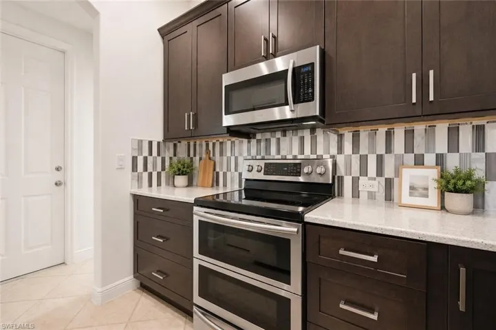 Kitchen featuring dark wood cabinetry with brushed nickel hardware, white speckled countertops, a mosaic tile backsplash, stainless steel microwave, and stainless steel electric range