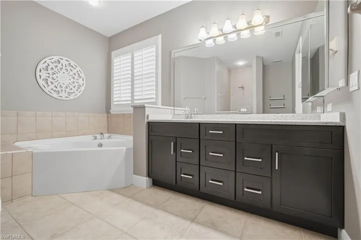 Bathroom featuring a corner soaking tub with tiled surround, plantation shutters, and a dark wood-finish vanity with a light-colored countertop and a wide mirror