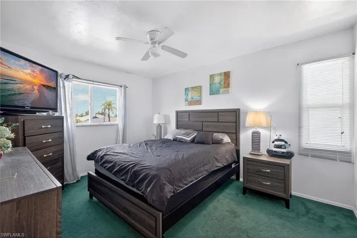 Room featuring crisp white walls, green carpet flooring, a window with horizontal blinds, a ceiling fan, and a second window with sheer curtains