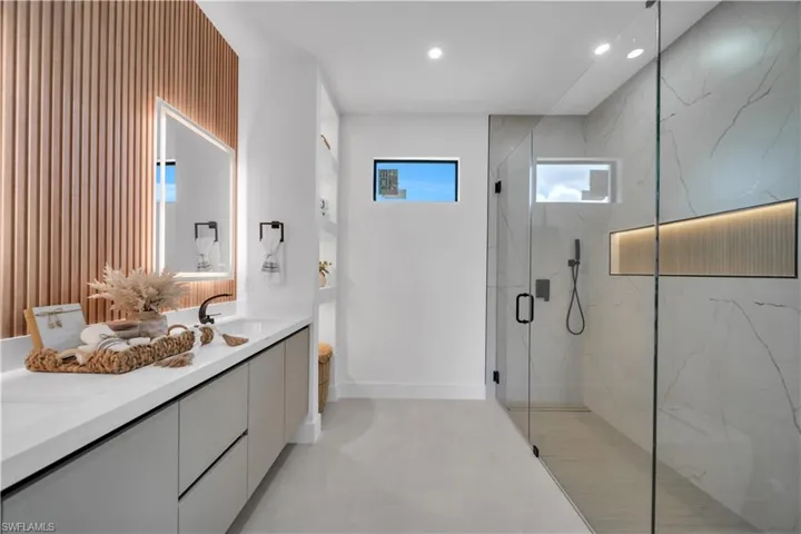 Contemporary bathroom featuring a spacious glass-enclosed shower with a recessed niche, a dual vanity with a white countertop and integrated sinks, a back-lit mirror, and a decorative wood-slat accent wall