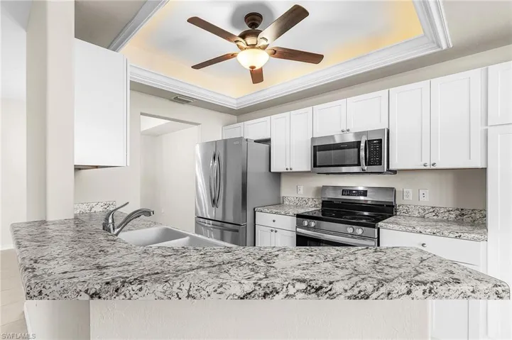Kitchen featuring stainless steel appliances, white cabinets, a ceiling fan, light stone countertops, and a raised ceiling