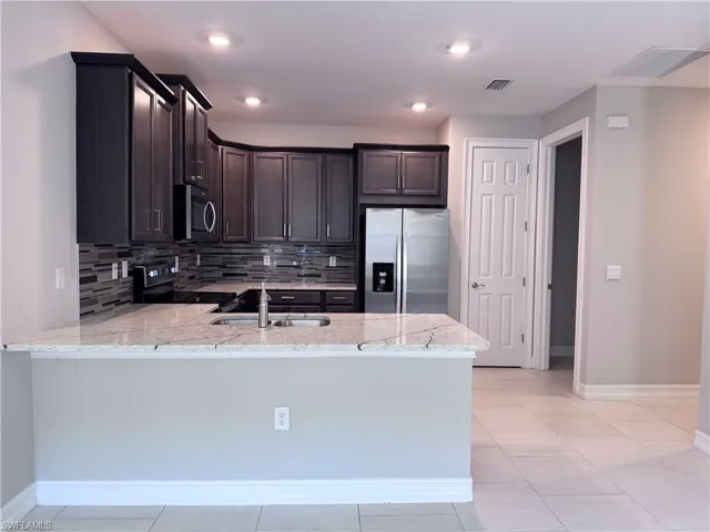 Kitchen with backsplash, appliances with stainless steel finishes, dark brown cabinets, light tile patterned floors, and light stone counters