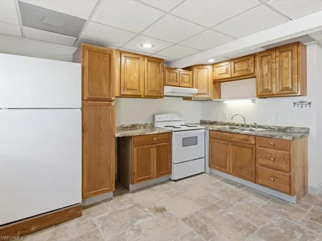 Kitchen featuring white appliances, a paneled ceiling, brown cabinets, under cabinet range hood, and light stone finish floors