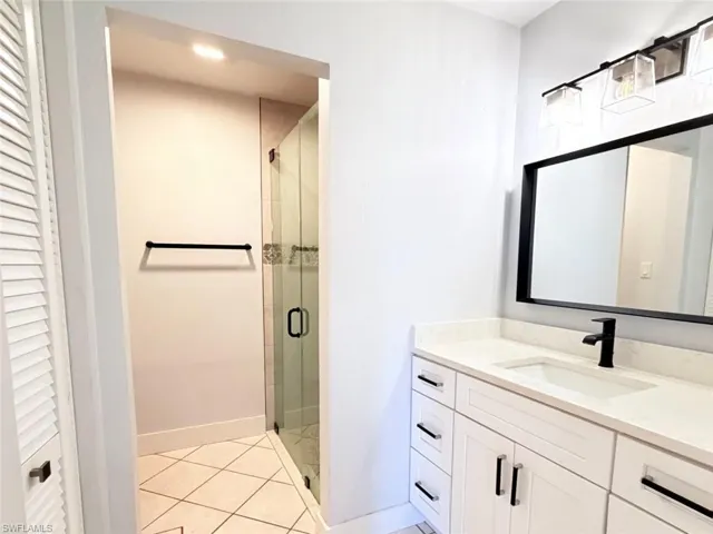 White cabinetry with dark hardware, quartz countertop, rectangular undermount sink, matte black faucet, and glass-enclosed shower with tiled flooring