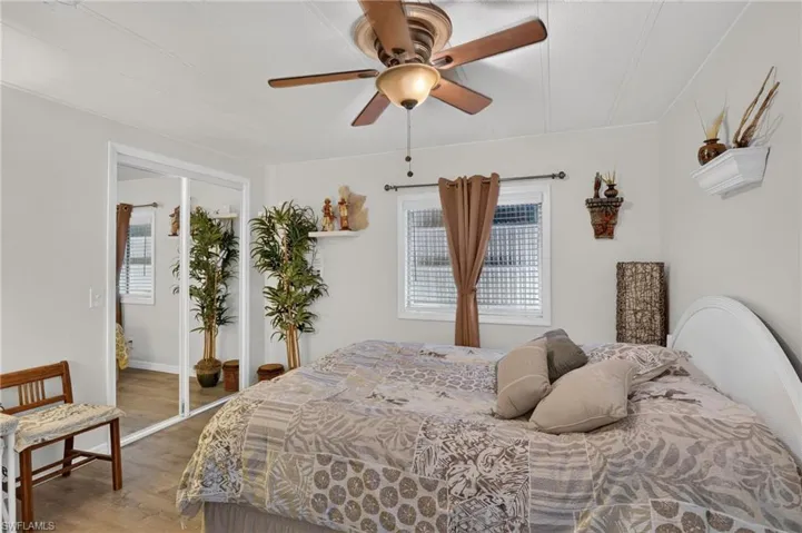 Wood-finish flooring, ceiling fan with integrated lighting, mirrored sliding closet doors, white paneled headboard, and a window with blinds