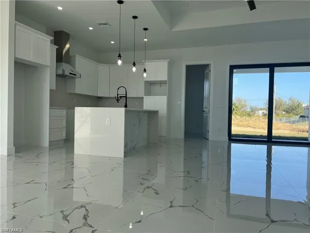 Kitchen featuring visible vents, white cabinets, wall chimney range hood, a center island with sink, and decorative light fixtures