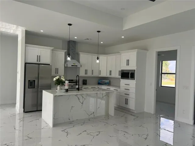 Kitchen featuring stainless steel appliances, hanging light fixtures, white cabinets, wall chimney range hood, and an island with sink