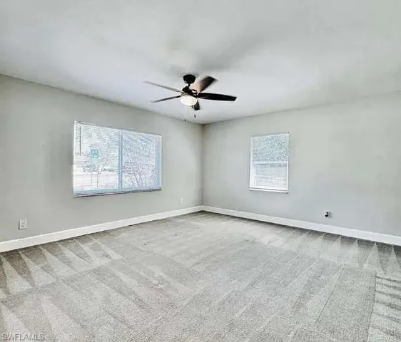 Expansive room featuring light gray walls, large windows with blinds, a ceiling fan with integrated lighting, and neutral-toned carpeting