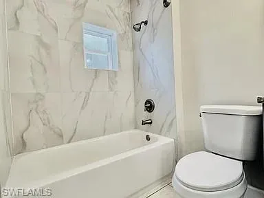 White bathtub featuring a window-mounted shower head and handheld sprayer, surrounded by large format wall tile