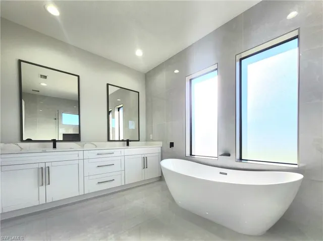 Full bath featuring healthy amount of natural light, a freestanding bath, double vanity, and light marble finish floors