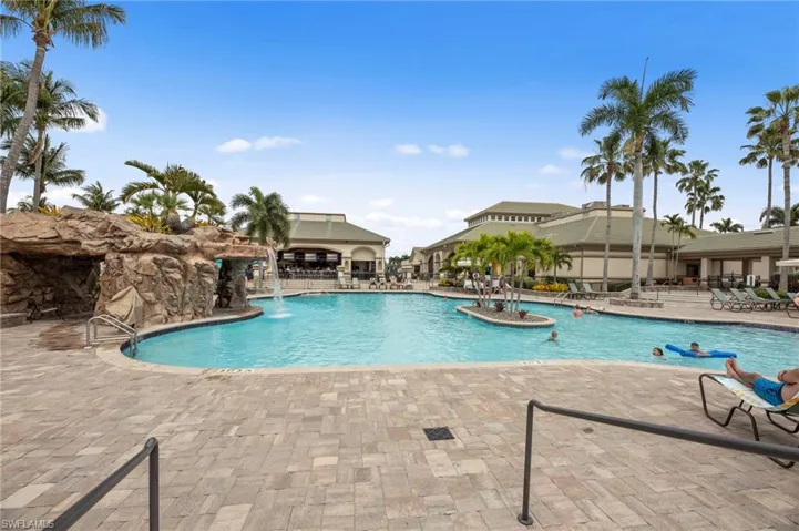Resort-style swimming pool featuring a rock grotto with waterfall, lush palm trees, and an expansive paver deck