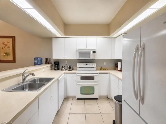 U-shaped kitchen featuring white cabinetry, light-toned countertops, a double basin stainless steel sink, and white appliances