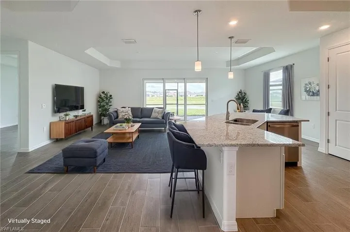 Kitchen with a tray ceiling, light stone counters, an island with sink, hanging light fixtures, and dark wood-type flooring
