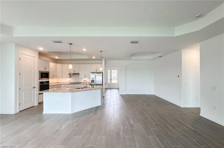 Kitchen with a tray ceiling, hanging light fixtures, a kitchen island with sink, wood finish floors, and light stone countertops