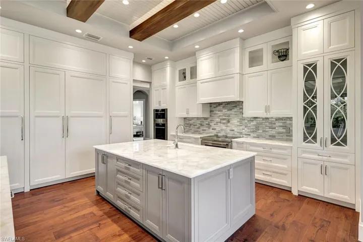 Kitchen with light stone countertops, a center island with sink, white cabinets, and beamed ceiling