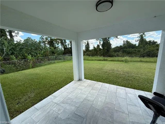 Covered concrete patio with neutral-toned tiling, featuring expansive grass lawn, chain-link perimeter fencing, and mature treeline