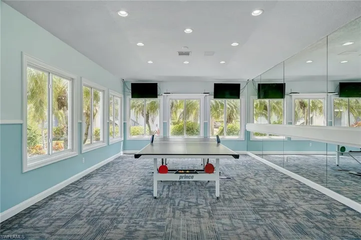 Spacious recreation room featuring multiple windows, recessed lighting, wall-mounted television screens, a full-wall mirror, and patterned carpet flooring