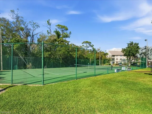Hard surface tennis court with perimeter fencing, surrounded by mature trees and a grassy expanse