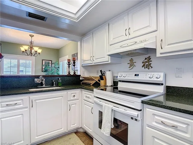 Kitchen with white range with electric cooktop, white cabinets, under cabinet range hood, a chandelier, and dark stone countertops