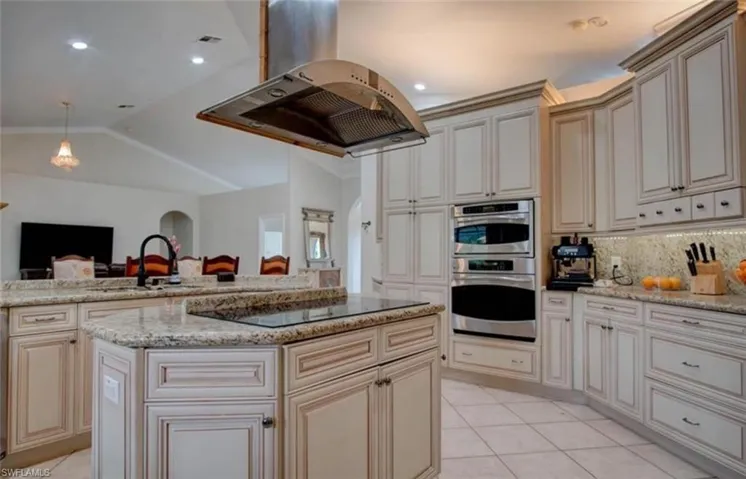 Kitchen featuring an expansive island with cooktop, granite countertops, custom cabinetry, stainless steel double wall ovens, and built-in range hood
