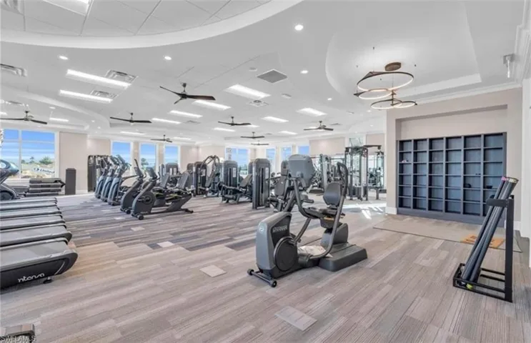 Spacious fitness center featuring varied exercise equipment, a large built-in shelving unit, abundant natural light from multiple windows, modern ceiling fans, and contemporary lighting fixtures