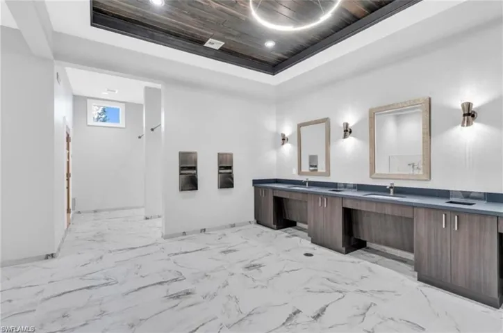 Spacious restroom featuring marble-look flooring, multiple vanity stations with integral sinks, dark wood-finish cabinetry, and a textured wood-plank ceiling with recessed lighting