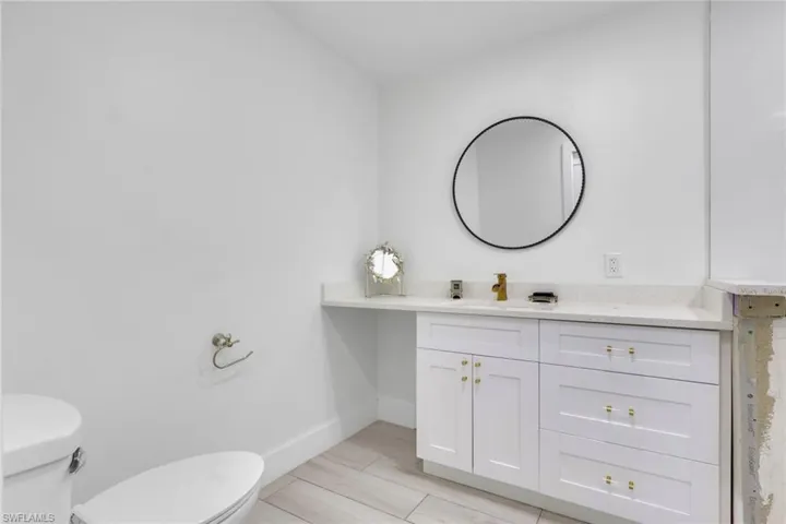 Bathroom featuring a white vanity with gold hardware, a circular wall-mounted mirror, a gold-finish faucet, a white countertop, and wood-finish tile flooring