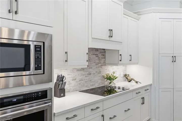 Shaker-style white cabinetry with brushed metal pulls, stone-composite countertops, horizontal-patterned stone tile backsplash, stainless steel built-in oven and microwave, and glass cooktop