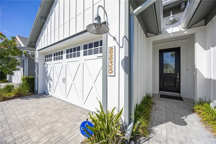White vertical board-and-batten siding, carriage-style garage door with glass panes, paver driveway and entryway, dark-finished front entry door, and exterior gooseneck lighting