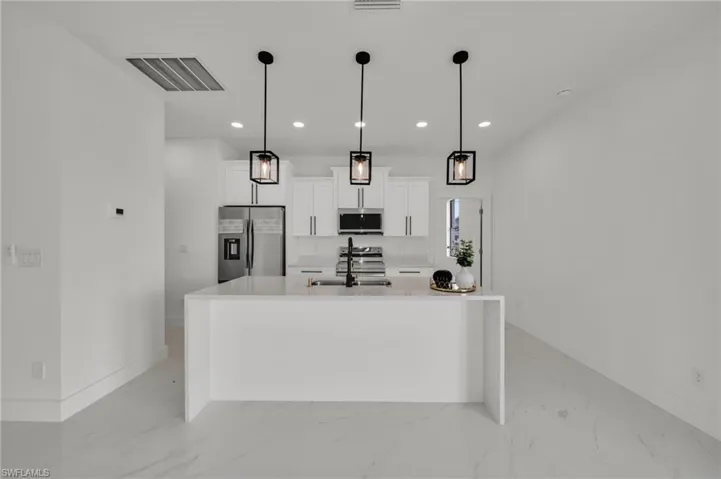 Kitchen with stainless steel appliances, light stone counters, white cabinetry, decorative light fixtures, and a kitchen island with sink