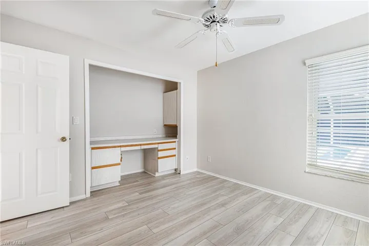 Light wood-finish flooring, built-in desk with cabinetry, window with blinds, and a ceiling fan