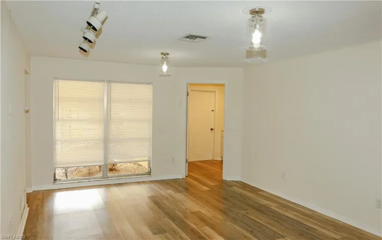 Spacious room with wood-finish flooring, neutral-toned walls, a large window with privacy blinds, and a combination of track lighting and flush-mount fixtures