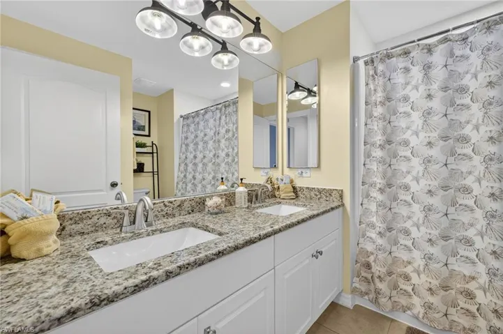 Guest bathroom with double vanity, tile-patterned flooring, chandelier accent lighting, and shower with curtain — stylishly updated in this Cape Coral furnished home.