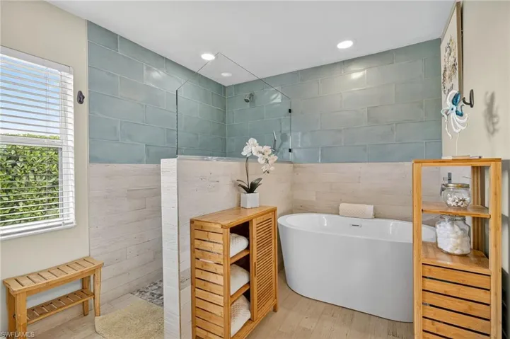 Primary bathroom with full tile walls, walk-in shower, freestanding soaking tub, wood-finish floors, and recessed lighting — elegantly appointed in this Cape Coral home.