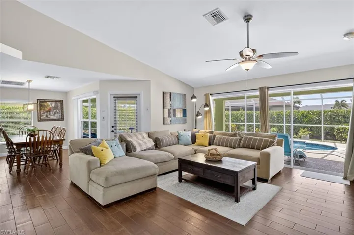 Bright and open living room with rich dark wood-style flooring, soaring vaulted ceilings, abundant natural light, and ceiling fan — ideal for entertaining in this furnished Cape Coral pool home.