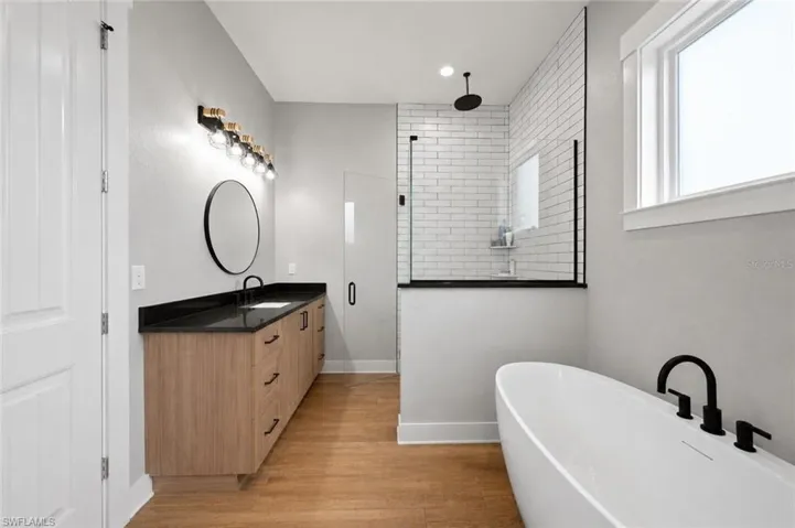 Modern bathroom featuring a freestanding tub with matte black fixtures, a walk-in shower with subway tile, a wood-finish double vanity with a dark countertop and round mirror, and wood-look flooring