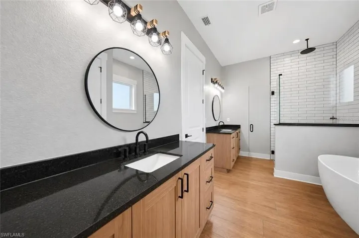 Bathroom featuring dual light wood-finish vanities with black countertops, a freestanding bathtub, a walk-in shower with subway tile, and wood-finish flooring