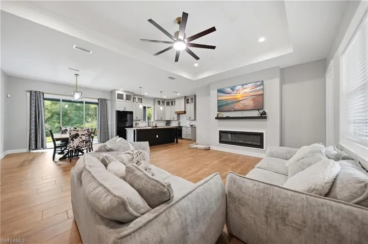 Open-concept living area featuring wood-finish flooring, a tray ceiling with recessed lighting, an electric fireplace with a floating mantel, and abundant natural light from multiple windows