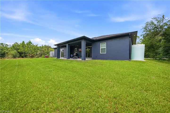 Expansive grassy backyard featuring a modern stucco exterior with a dark roofline, covered patio, and a water storage tank