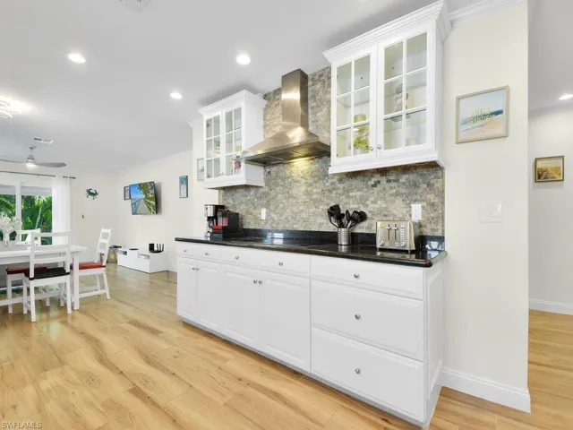 Kitchen with glass insert cabinets, wall chimney range hood, white cabinetry, light wood-type flooring, and recessed lighting