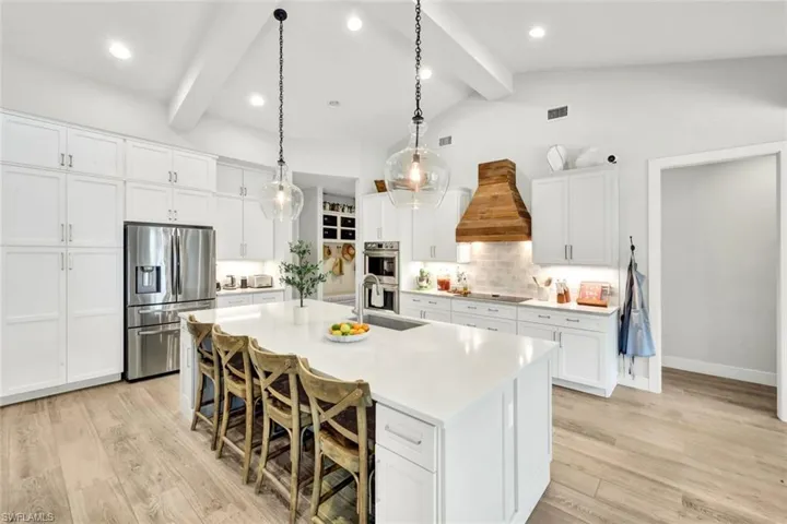Kitchen featuring appliances with stainless steel finishes, white cabinets, decorative light fixtures, custom range hood, and a kitchen bar