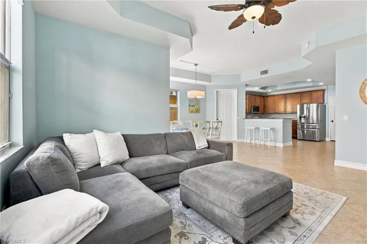 Open-concept living space featuring high tray ceilings, ceramic tile flooring, a multi-blade ceiling fan, and a kitchen equipped with stainless steel appliances and wood cabinetry