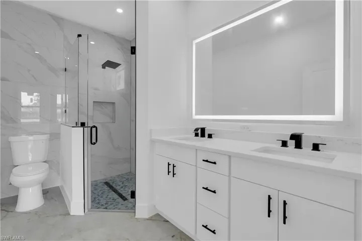 Contemporary bathroom featuring a dual vanity with white cabinetry and matte black hardware, a back-lit mirror, a glass-enclosed shower with marble-look wall tile and a rainfall showerhead, and a built-in shower niche