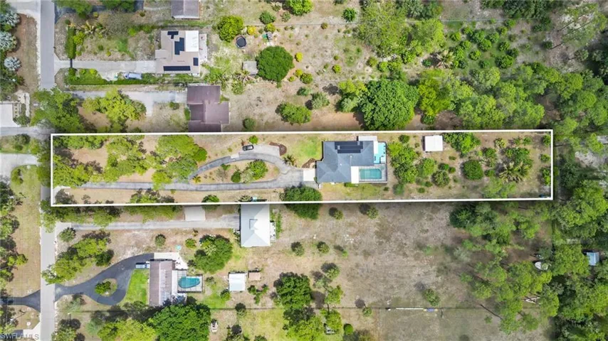 Expansive property featuring a winding driveway, a single-story residence with a dark-toned roof, a rectangular swimming pool, and a mix of mature trees and natural landscaping