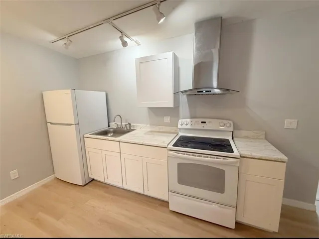 Kitchen featuring light wood finished floors, baseboards, white appliances, a sink, and wall chimney range hood