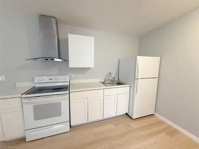 Kitchen featuring wall chimney range hood, a sink, white cabinetry, and white appliances