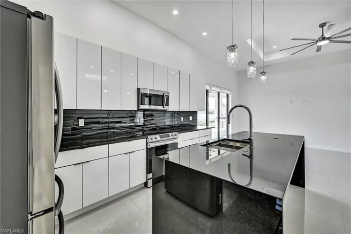 Modern kitchen featuring white high-gloss cabinetry, black granite countertops, stainless steel appliances, and a central island with an integrated sink and gooseneck faucet
