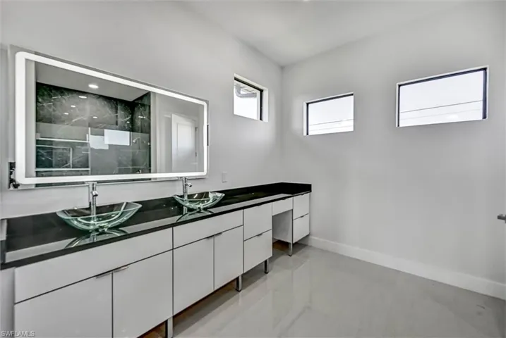 Contemporary bathroom featuring a dual vanity with vessel sinks, an illuminated mirror, dark stone countertops, and light-toned floor tiles