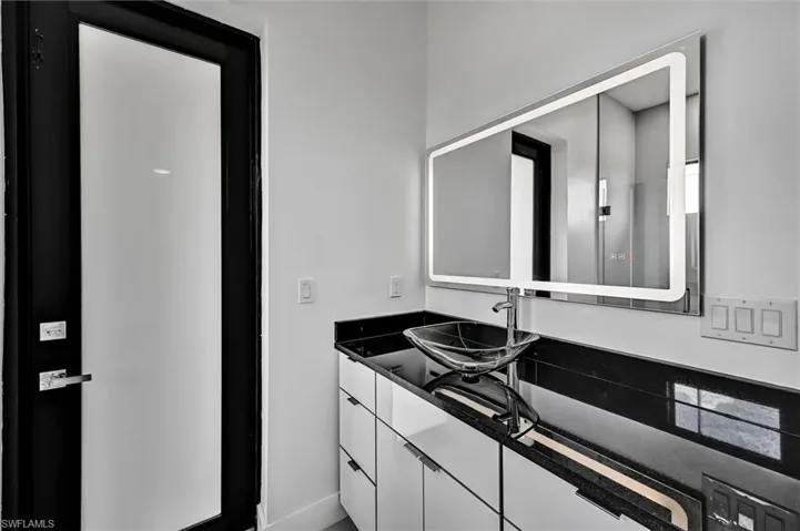 Modern vanity featuring a black countertop, clear vessel sink, and a large illuminated mirror